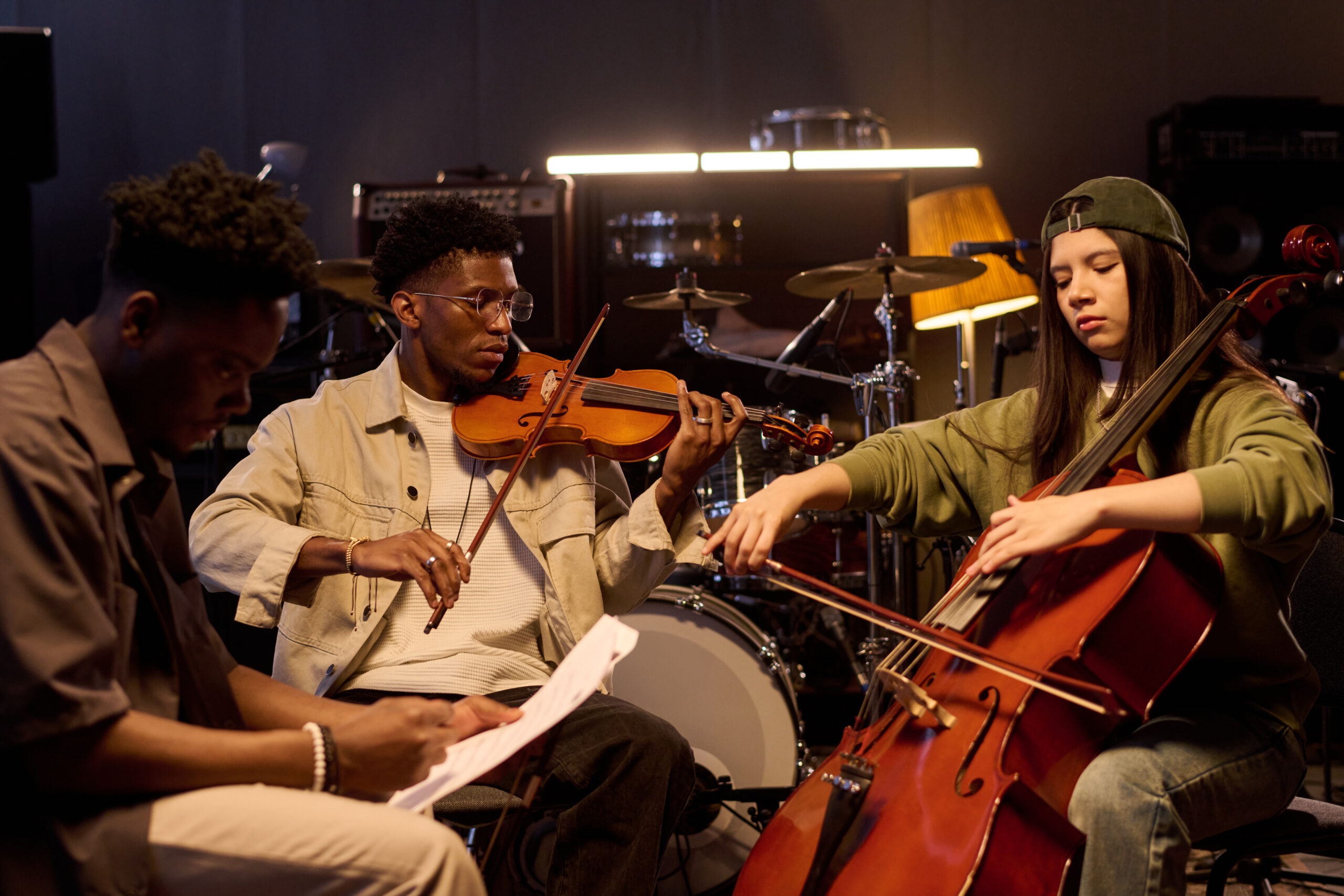 Black young adult man playing violin next to Latin young adult woman playing cello while sitting in music studio with musical instruments, another Black young adult man reading sheet music